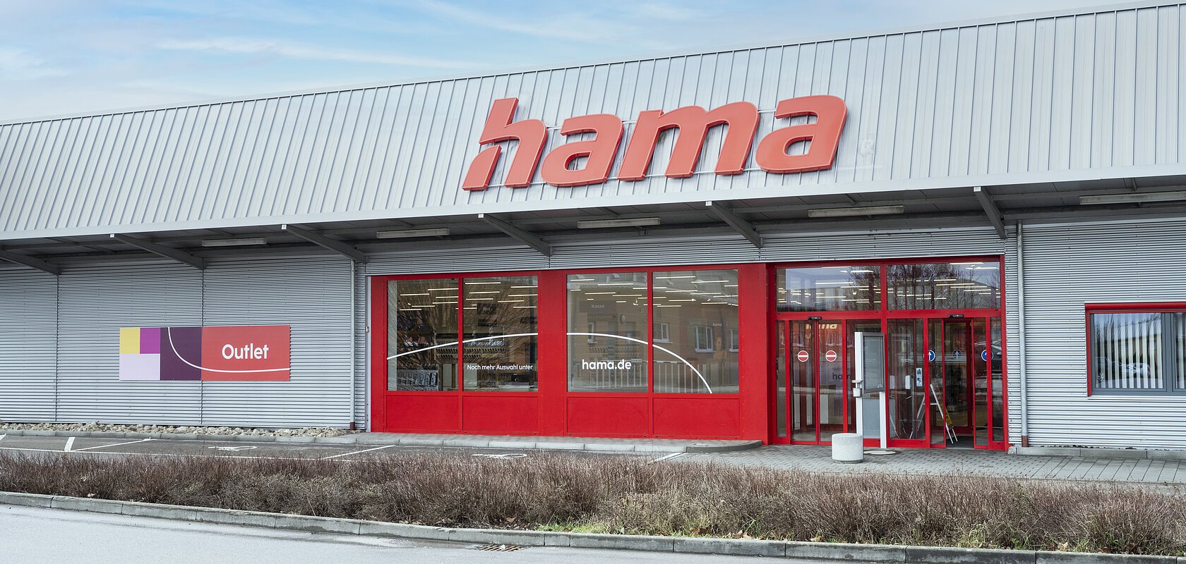 Graue Fassade eines Outlet-Geschäfts mit rotem Schriftzug "Hama", roten Eingangstüren und roten Fensterumrandungen, bei bewölktem Himmel. Vor dem Geschäft eine Hecke entlang einer Straße.