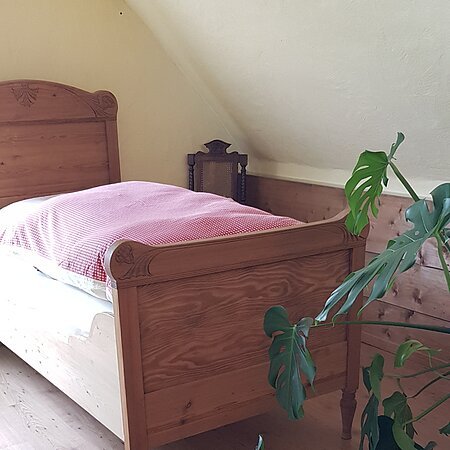 Holzbett mit roter Decke drauf in einem Zimmer mit schräger gelbgestrichener Decke, Holzboden und Holzverkleidung an der Wand hinter dem Bett, grüner Kommode und grüner Pflanze im Vordergrund.