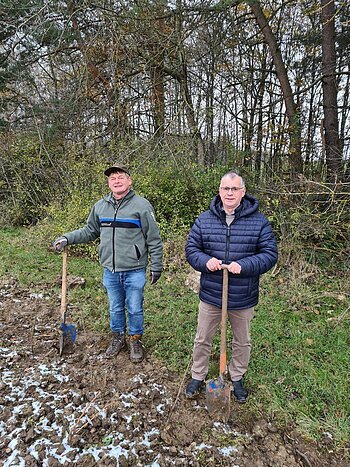 Ersatzaufforstung nach Rodung Zwei Männer stehen blickend in die Kamera mit jeweils einem Spaten auf einem teilweise verschneiten Acker vor Büschen und Bäumen.