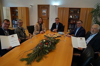 Sechs Personen sitzen an einem Tisch und blicken in die Kamera, zwei davon halten eine Mappe mit einer Urkunde in die Kamera. Sektgläser, ein Teller mit belegten Broten und Dekoration aus Tannenzweigen in der Mitte des Tisches. Im Hintergrund Holzschränke mit Glasscheiben in einem Raum.