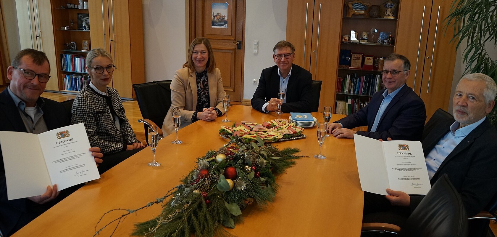 Sechs Personen sitzen an einem Tisch und blicken in die Kamera, zwei davon halten eine Mappe mit einer Urkunde in die Kamera. Sektgläser, ein Teller mit belegten Broten und Dekoration aus Tannenzweigen in der Mitte des Tisches. Im Hintergrund Holzschränke mit Glasscheiben in einem Raum.