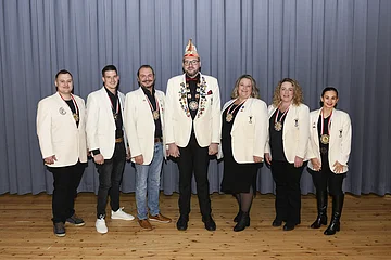 Sieben Personen in weißen Blazern mit Medaillen stehen vor einem grauen Vorhang auf Holzboden.