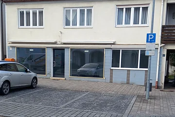 Zweistöckiges zweifarbiges Gebäude mit drei Fenstern oben und drei großen Schaufenstern unten. Ein parkendes Auto auf einem Parkplatz davor.