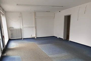 Leerer Raum mit blaugrauem Teppichboden und weißen Wänden, Heizkörper links hinten an der Wand und offener Tür rechts sichtbar.