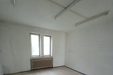 Leerer Raum mit weißen Wänden, zwei Fenstern, einem Heizkörper darunter und gefliestem Boden. An der Decke drei Leuchtröhren.