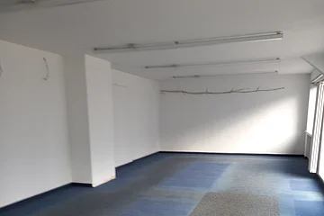 Leerer Raum mit weißen Wänden, blaugrauem Teppichboden und mehreren Deckenleuchten, Fenster rechts.