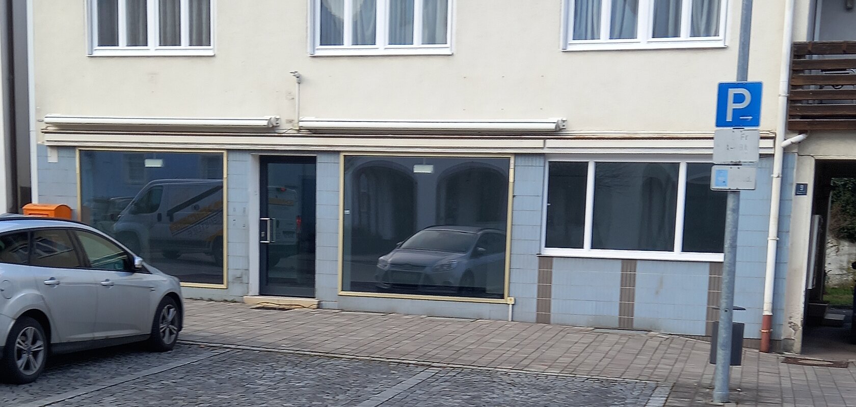 Zweistöckiges zweifarbiges Gebäude mit drei Fenstern oben und drei großen Schaufenstern unten. Ein parkendes Auto auf einem Parkplatz davor.