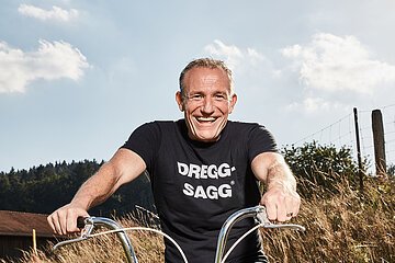 Mann mit schwarzem T-Shirt mit der weißen Aufschrift "DREGG-SAGG" und Jeans sitzt lachend auf orangefarbenem Fahrrad auf Feldweg bei Sonnenschein