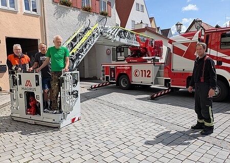 Drei Männer in ziviler Kleidung stehen in einem Feuerwehrkorb vor Wohnhäusern und Torbogen, ein Mann in Feuerwehr-Uniform steht rechts daneben, im Hintergrund ein Feuerwehrfahrzeug mit ausgefahrener Drehleiter, bei blauem Himmel.