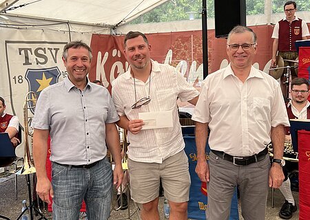 Drei Männer stehen nebeneinander vor einem Zelt mit Musikern im Hintergrund, einer hält einen Briefumschlag. Hinter ihnen ein Banner mit Aufschrift TSV 1895 Monheim sichtbar.