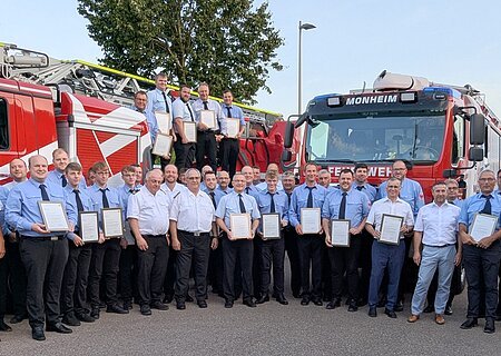 Gruppenfoto von Feuerwehrleuten in blauer Uniform in mehreren Reihen vor zwei roten Feuerwehrfahrzeugen, die meisten halten Urkunden, bei blauem Himmel.