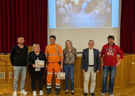 Sechs Personen stehen nebeneinander auf einer Bühne vor rotem Vorhang und Leinwand mit Jubelbild und Text. Es handelt sich um den Personalrat der Stadt Monheim und geehrte Jubilare der Stadt Monheim.