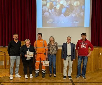 Sechs Personen stehen nebeneinander auf einer Bühne vor rotem Vorhang und Leinwand mit Jubelbild und Text. Es handelt sich um den Personalrat der Stadt Monheim und geehrte Jubilare der Stadt Monheim.
