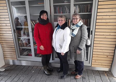Drei Frauen in Winterkleidung stehen vor einem Bücherschrank mit Glastüren links und rechts am Schrank eine Holzfassade.