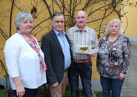 Vier Personen stehen nebeneinander vor einer gelben Wand mit kahlen Ästen, eine Person hält ein verpacktes Geschenk.