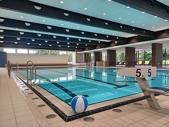 Innenaufnahme des leeren Schwimmbads - Jurabad Monheim. Zu sehen ist ein Schwimmbecken gefüllt mit strahlendem Wasser, ein Sprungbrett und ein weiß-blauer Wasserball liegt dekorativ im Vordergrund. Im Hintergrund des Hallenbads sind große Fenster zu sehen.