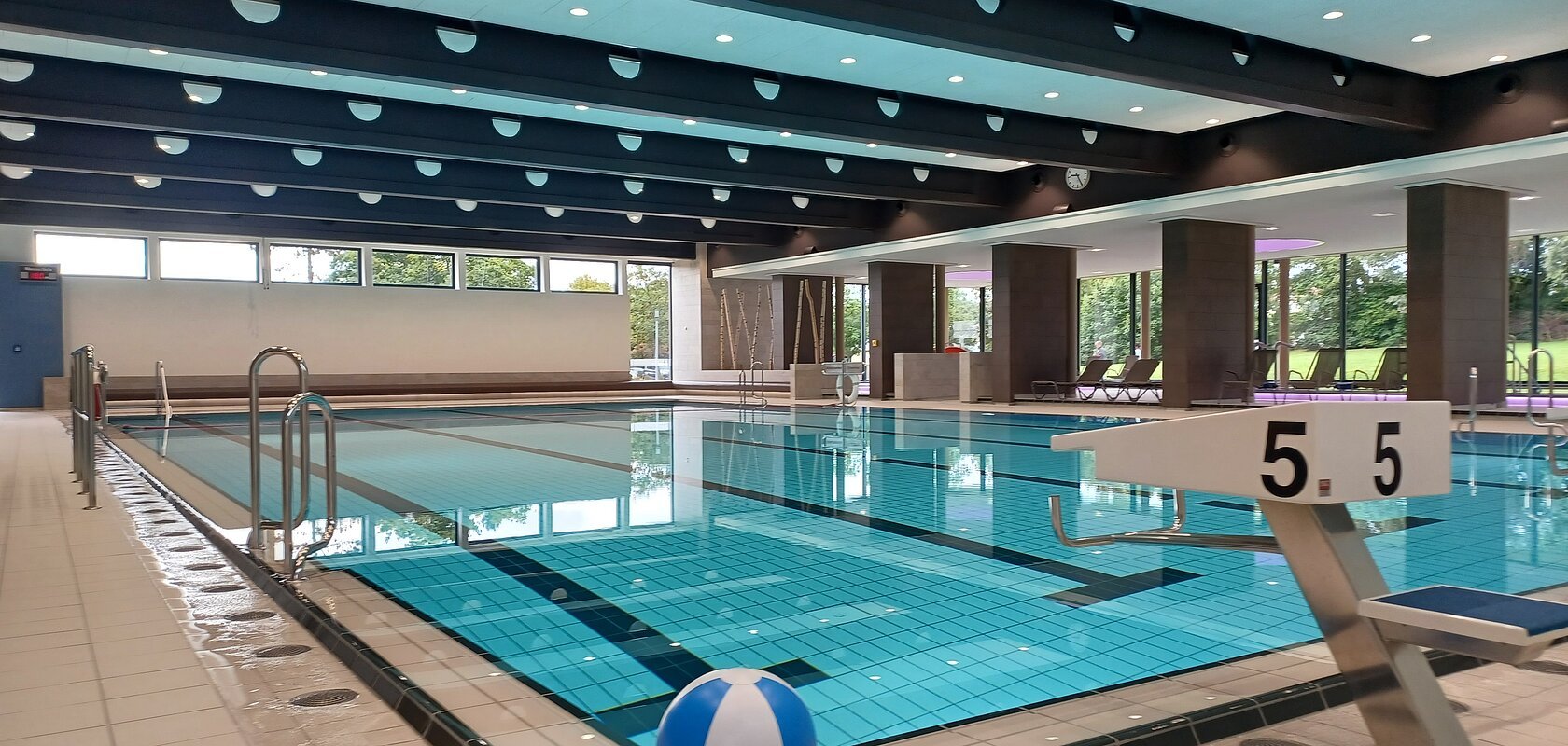 Innenaufnahme des leeren Schwimmbads - Jurabad Monheim. Zu sehen ist ein Schwimmbecken gefüllt mit strahlendem Wasser, ein Sprungbrett und ein weiß-blauer Wasserball liegt dekorativ im Vordergrund. Im Hintergrund des Hallenbads sind große Fenster zu sehen.