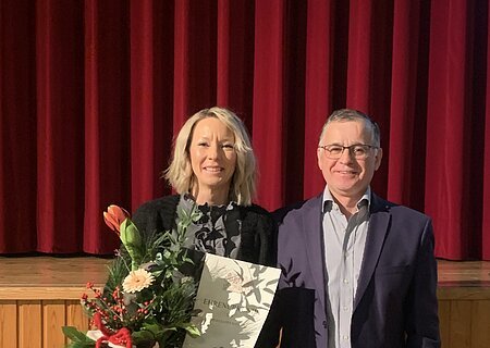 Zwei Personen vor rotem Bühnenvorhang, eine hält Blumenstrauß und Urkunde, beide lächeln in die Kamera blickend.