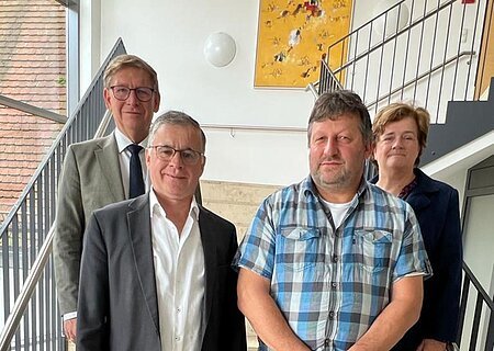 Vier Personen stehen blickend in die Kamera auf einer Treppe in einem Gebäude mit schwarzem Geländer und großem Fenster. An der Wand hängt ein gelbes Gemälde.