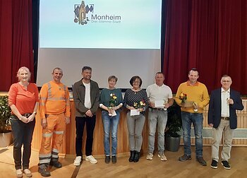 Acht Personen stehen nebeneinander vor einer Bühne mit rotem Vorhang und einer Leinwand mit Monheim-Logo, einige halten Blumen und Urkunden.