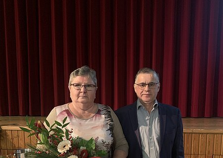 Zwei Personen vor rotem Bühnenvorhang, Frau hält Blumenstrauß, Mann trägt Anzug und Brille. Im Hintergrund ein gedeckter Tisch.