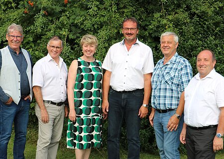 Sechs Personen stehen nebeneinander vor  grünen Bäumen, alle blicken in die Kamera und lächeln.