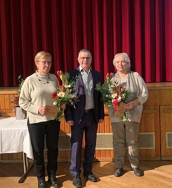 Drei Personen stehen vor rotem Bühnenvorhang, ein Mann steht in der Mitte, links und rechts zwei Frauen, die Blumensträuße und Umschläge in den Händen halten. Hinter ihnen ein Tisch mit weißer Decke.
