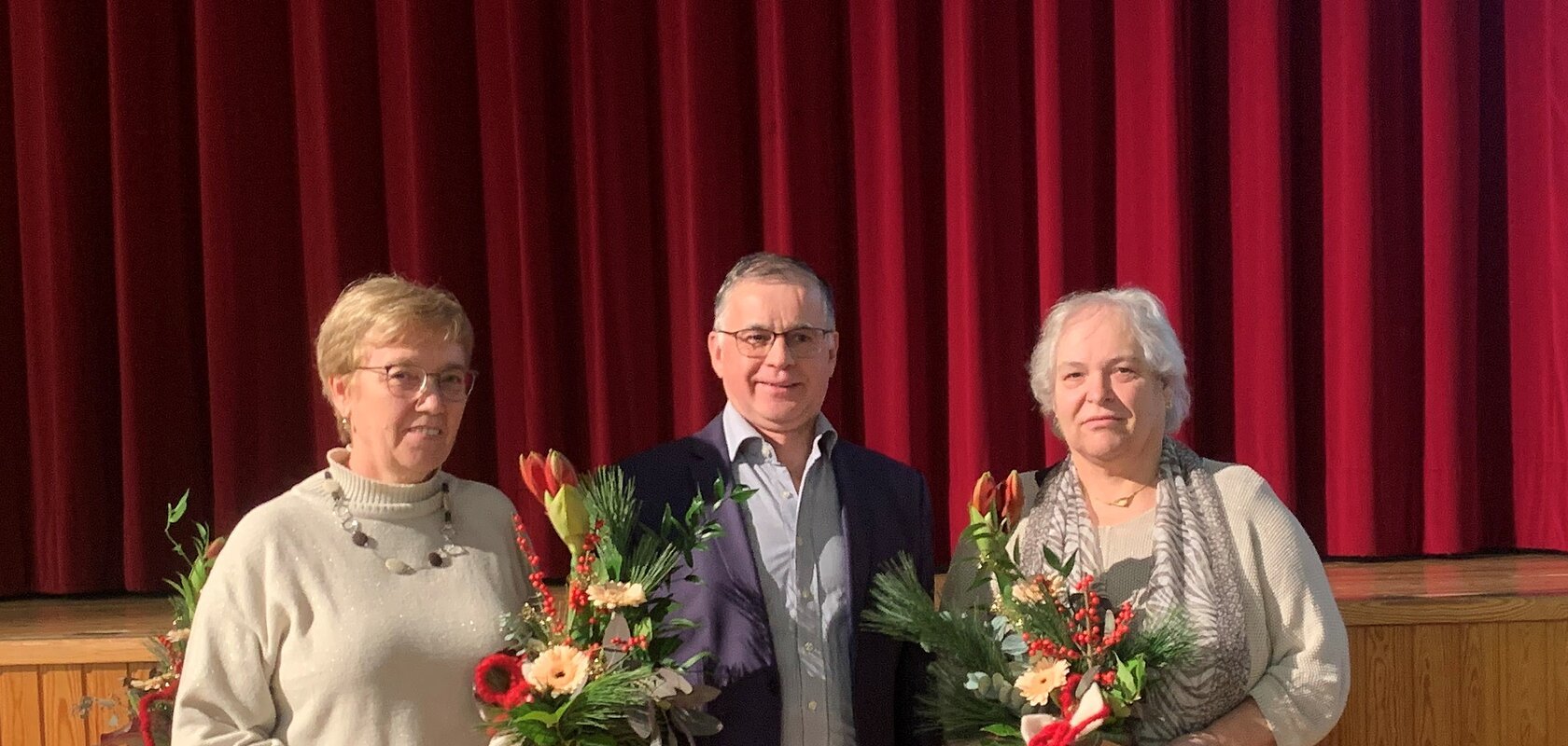 Drei Personen stehen vor rotem Bühnenvorhang, ein Mann steht in der Mitte, links und rechts zwei Frauen, die Blumensträuße und Umschläge in den Händen halten. Hinter ihnen ein Tisch mit weißer Decke.