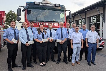 Elf Personen in Feuerwehruniform und zwei Personen in Hemd stehen vor einem roten Feuerwehrfahrzeug mit der Aufschrift „Feuerwehr Monheim“ auf einem gepflastertem Platz.