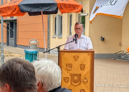 Mann in weißem Hemd mit Brille hält Rede am Rednerpult aus Holz mit 9 verschiedenen Wappen an der Front unter einem schwarz-orangem Sonnenschirm auf einem gepflastertem Platz. Davor zwei Personen mit schwarzen Kolping Monheim Shirts.