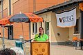 Frau im knalligem grünem T-Shirt spricht am Rednerpult aus Holz mit 9 verschiedenen Wappen unter einem Kolping-Schirm in orange-schwarz. Rechts ein Banner am Gebäude mit dem Schriftzug "Kolping" sichtbar, sowie orange Luftballons. Davor zwei Personen von Hinten mit Kolping-Monheim-Shirts.