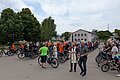 Große Gruppe von Radfahrern mit ihren Fahrrädern auf einem Parkplatz, ein Stadtpfarrer mit liturgischem Gewand segnet die Gruppe. Neben ihm ein weiterer Mann mit einem Behälter mit Weihwasser.