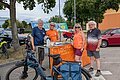 Vier Personen stehen an einem Infostand mit orangefarbenem ADFC-Banner und mehreren Fahrrädern im Hintergrund auf einem Parkplatz. Im Hintergrund Bäume und ein oranges Gebäude unter bewölktem Himmel.