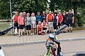 Gruppe von 15 Personen steht auf einem Gehweg vor Bäumen, im Vordergrund Fahrradlenker und Straße sichtbar.