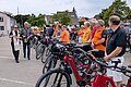 Rechts im Bild eine Gruppe von Menschen mit Fahrrädern auf einem Parkplatz, ein Stadtpfarrer mit liturgischem Gewand und ein weiterer mit einem Eimer gehen an ihnen vorbei und segnen die Radfahrer mit Weihwasser.