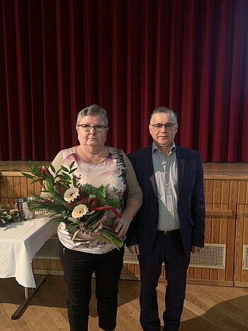Zwei Personen vor rotem Bühnenvorhang, Frau hält Blumenstrauß, Mann trägt Anzug und Brille. Im Hintergrund ein gedeckter Tisch.