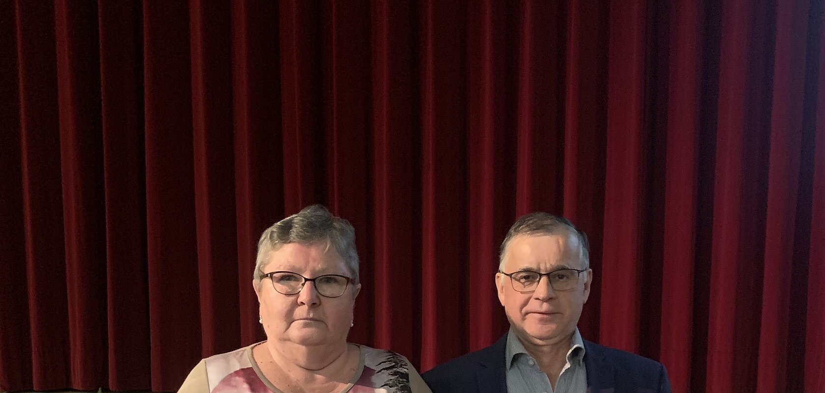 Zwei Personen vor rotem Bühnenvorhang, Frau hält Blumenstrauß, Mann trägt Anzug und Brille. Im Hintergrund ein gedeckter Tisch.