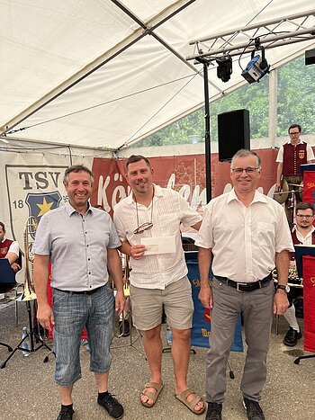Drei Männer stehen nebeneinander vor einem Zelt mit Musikern im Hintergrund, einer hält einen Briefumschlag. Hinter ihnen ein Banner mit Aufschrift TSV 1895 Monheim sichtbar.