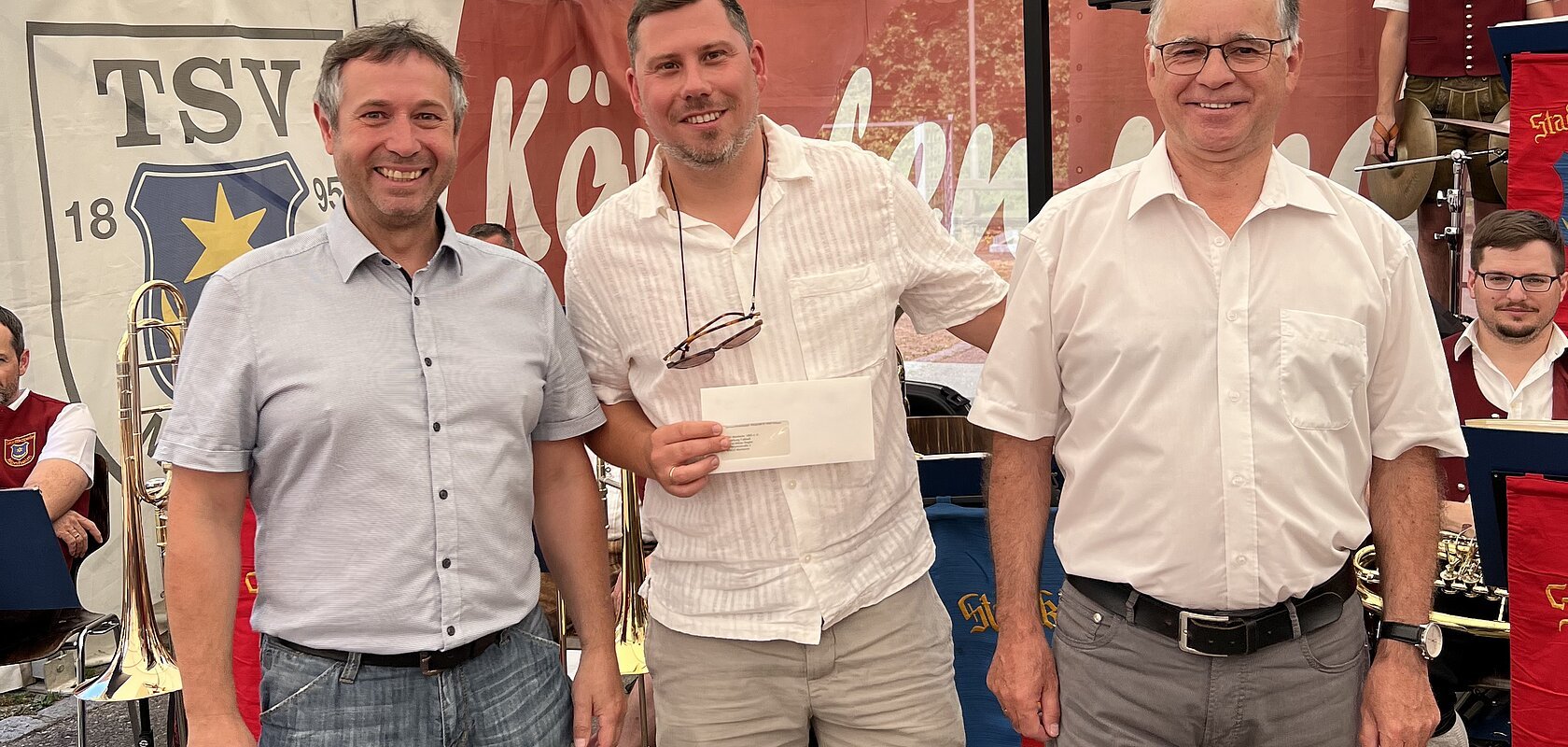 Drei Männer stehen nebeneinander vor einem Zelt mit Musikern im Hintergrund, einer hält einen Briefumschlag. Hinter ihnen ein Banner mit Aufschrift TSV 1895 Monheim sichtbar.