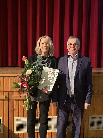 Zwei Personen vor rotem Bühnenvorhang, eine hält Blumenstrauß und Urkunde, beide lächeln in die Kamera blickend.