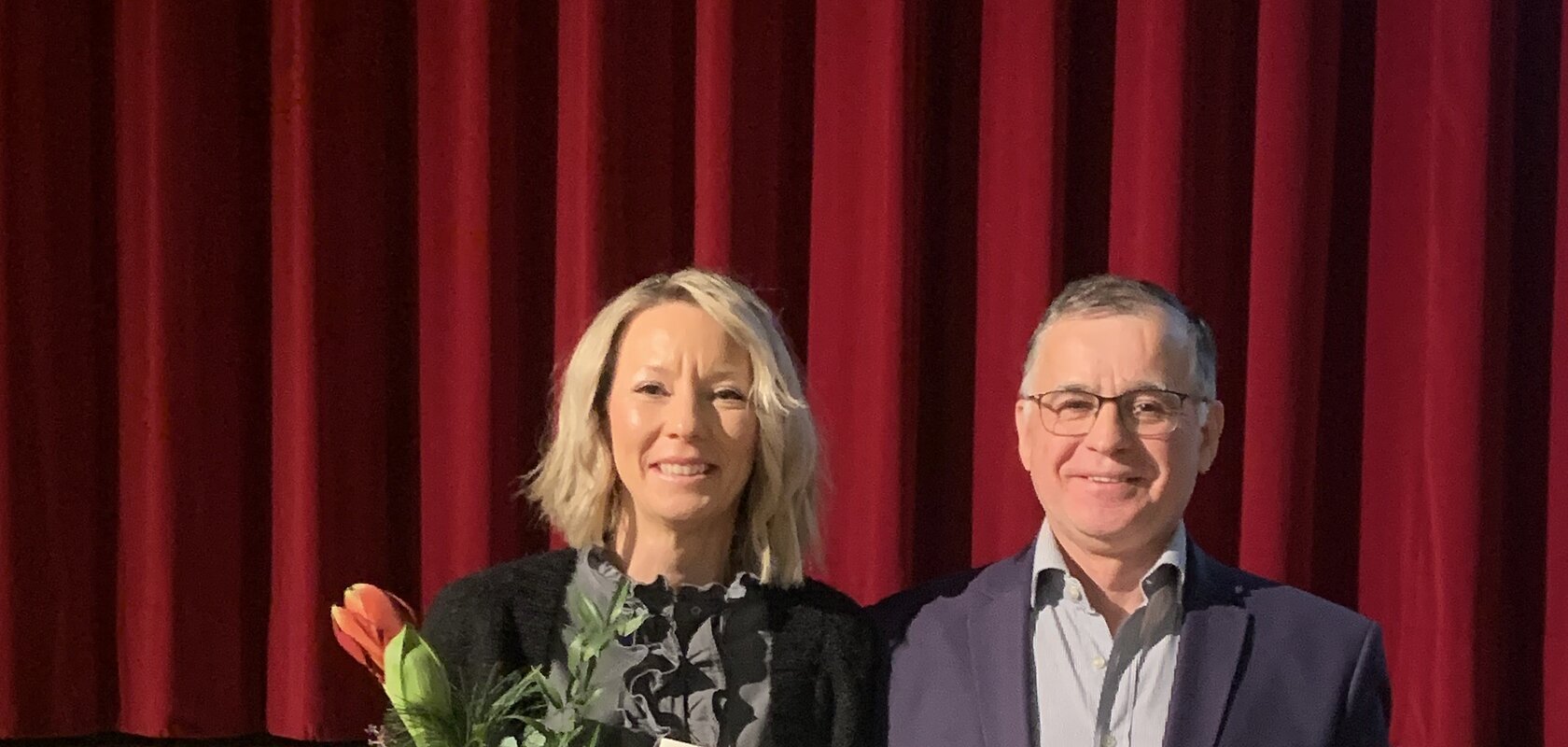 Zwei Personen vor rotem Bühnenvorhang, eine hält Blumenstrauß und Urkunde, beide lächeln in die Kamera blickend.