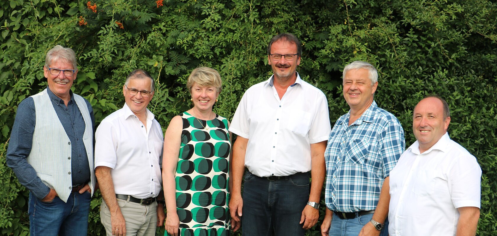 Sechs Personen stehen nebeneinander vor  grünen Bäumen, alle blicken in die Kamera und lächeln.