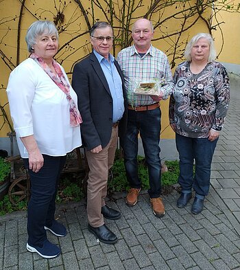 Vier Personen stehen nebeneinander vor einer gelben Wand mit kahlen Ästen, eine Person hält ein verpacktes Geschenk.