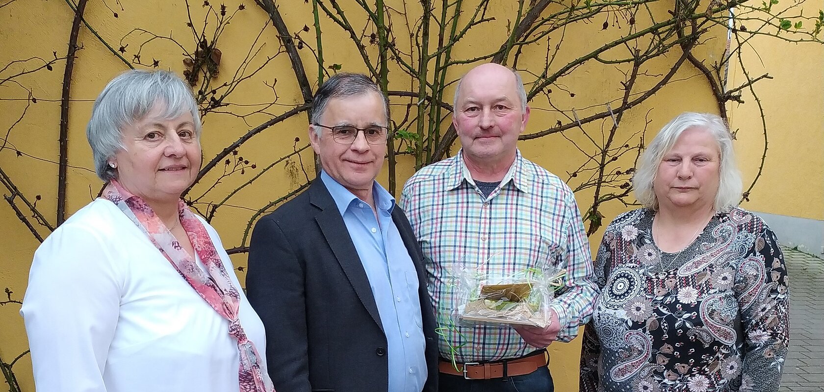 Vier Personen stehen nebeneinander vor einer gelben Wand mit kahlen Ästen, eine Person hält ein verpacktes Geschenk.