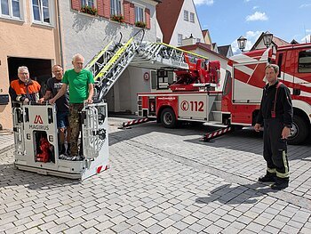 Drei Männer in ziviler Kleidung stehen in einem Feuerwehrkorb vor Wohnhäusern und Torbogen, ein Mann in Feuerwehr-Uniform steht rechts daneben, im Hintergrund ein Feuerwehrfahrzeug mit ausgefahrener Drehleiter, bei blauem Himmel.