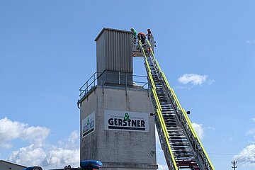 Feuerwehrfahrzeug mit ausgefahrener Drehleiter vor einem hohen Gebäude mit Gerstner-Schildern und drei Personen oben auf der Leiter, bei blauem Himmel.