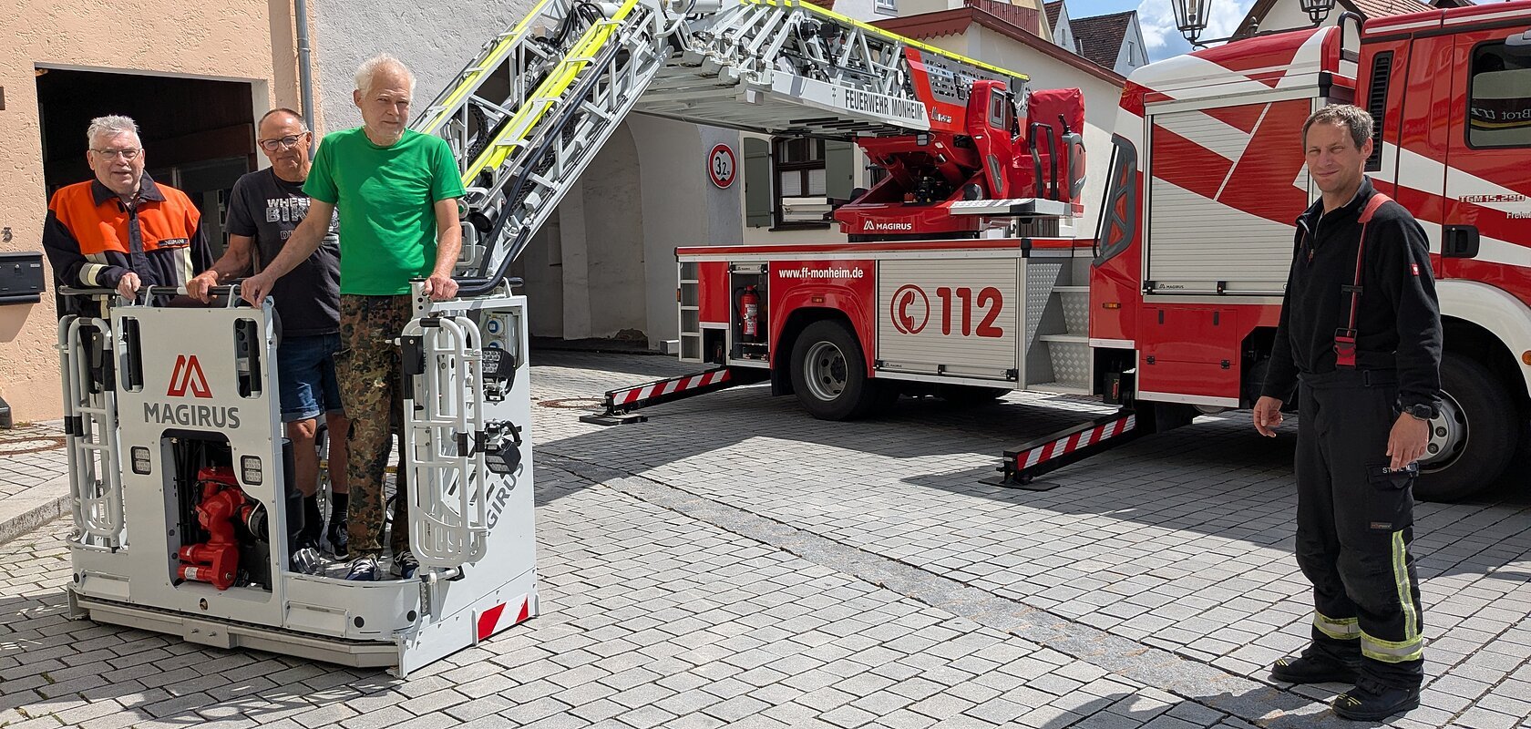 Drei Männer in ziviler Kleidung stehen in einem Feuerwehrkorb vor Wohnhäusern und Torbogen, ein Mann in Feuerwehr-Uniform steht rechts daneben, im Hintergrund ein Feuerwehrfahrzeug mit ausgefahrener Drehleiter, bei blauem Himmel.