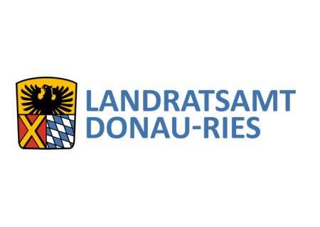 Wappen des Landratsamts DONAU-RIES: auf dem Wappen oben ein schwarzer Adler auf gelbem Hintergrund. darunter links ein gelbes Kreuz auf rotem Hintergrund und rechts davon bayerisches Rautenmuster in weiß-blau. Rechts neben dem Wappen der Text: Landratsamt Donau-Ries.