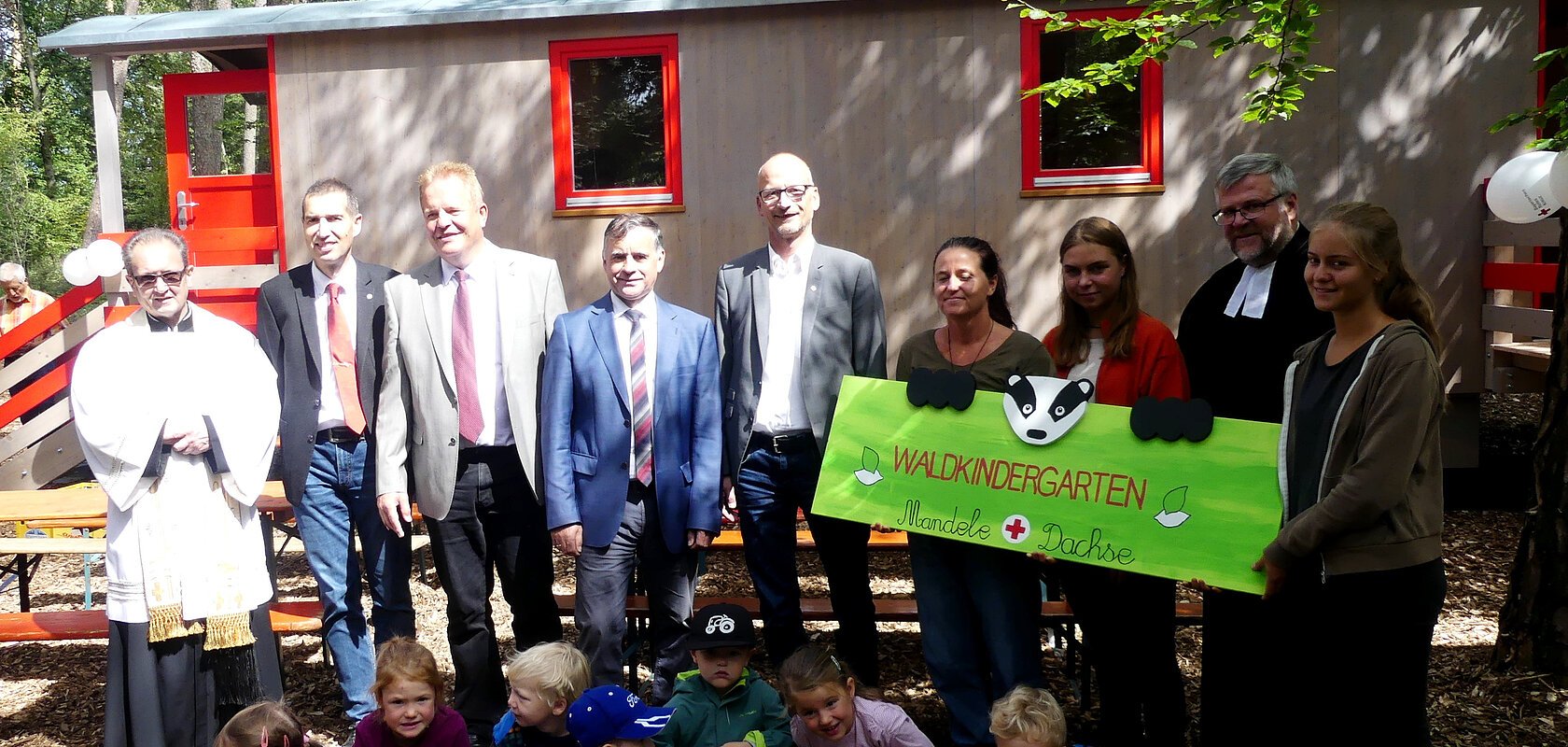 Eine Gruppe von Erwachsenen und Kindern steht bzw. kniet vor einem Bauwagen im Wald, drei Personen halten ein gestaltetes Schild mit der Aufschrift "Waldkindergarten Mandele Dachse" in den Händen.