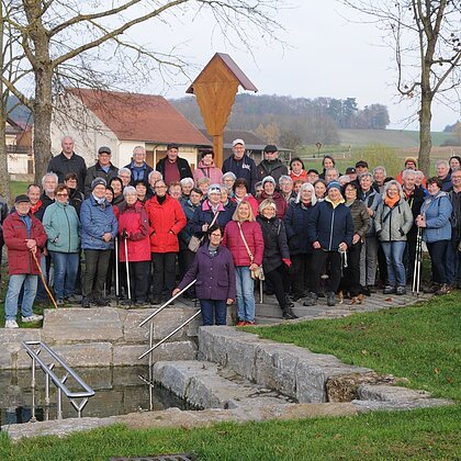 Große Gruppe älterer Menschen steht im Park vor einem kleinen Wasserbecken mit Treppe und Geländer. Monatliche Wanderung für Senioren vom Verein Soziales Miteinander